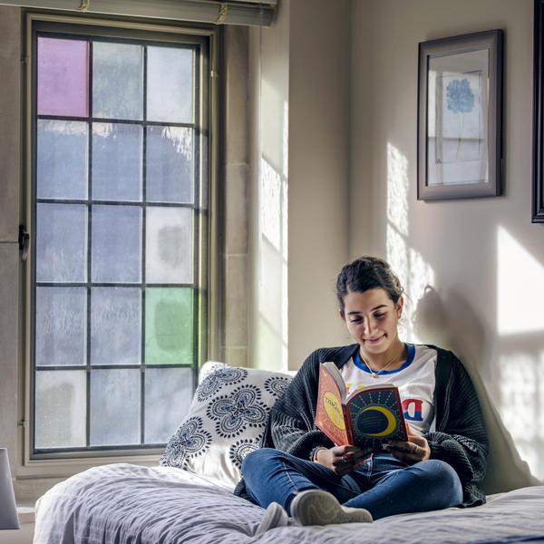 一位年轻女子坐在床上，借着彩色玻璃窗的灯光看书.